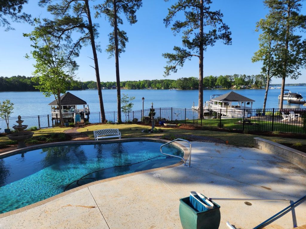 Backyard swimming pool with a lake view, surrounded by trees, fencing, and docks on a sunny day.