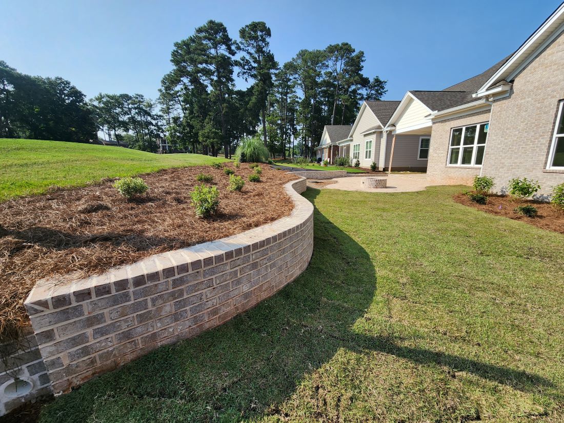 A landscaped yard featuring a curved brick retaining wall with a mulched garden bed on top, containing small shrubs and plants in Lexington, SC