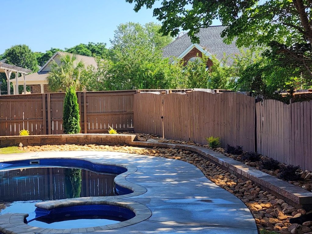 Backyard landscaping around an in-ground pool with a wooden fence and young plants.