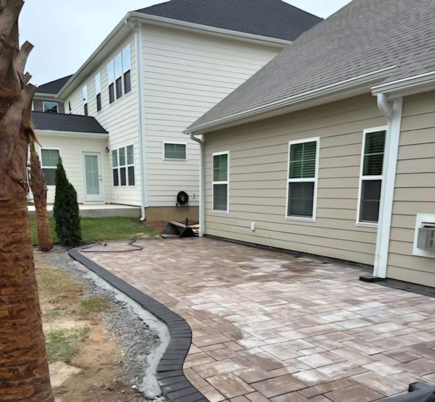 Back patio area under construction with stone pavers partially installed beside a beige siding house.