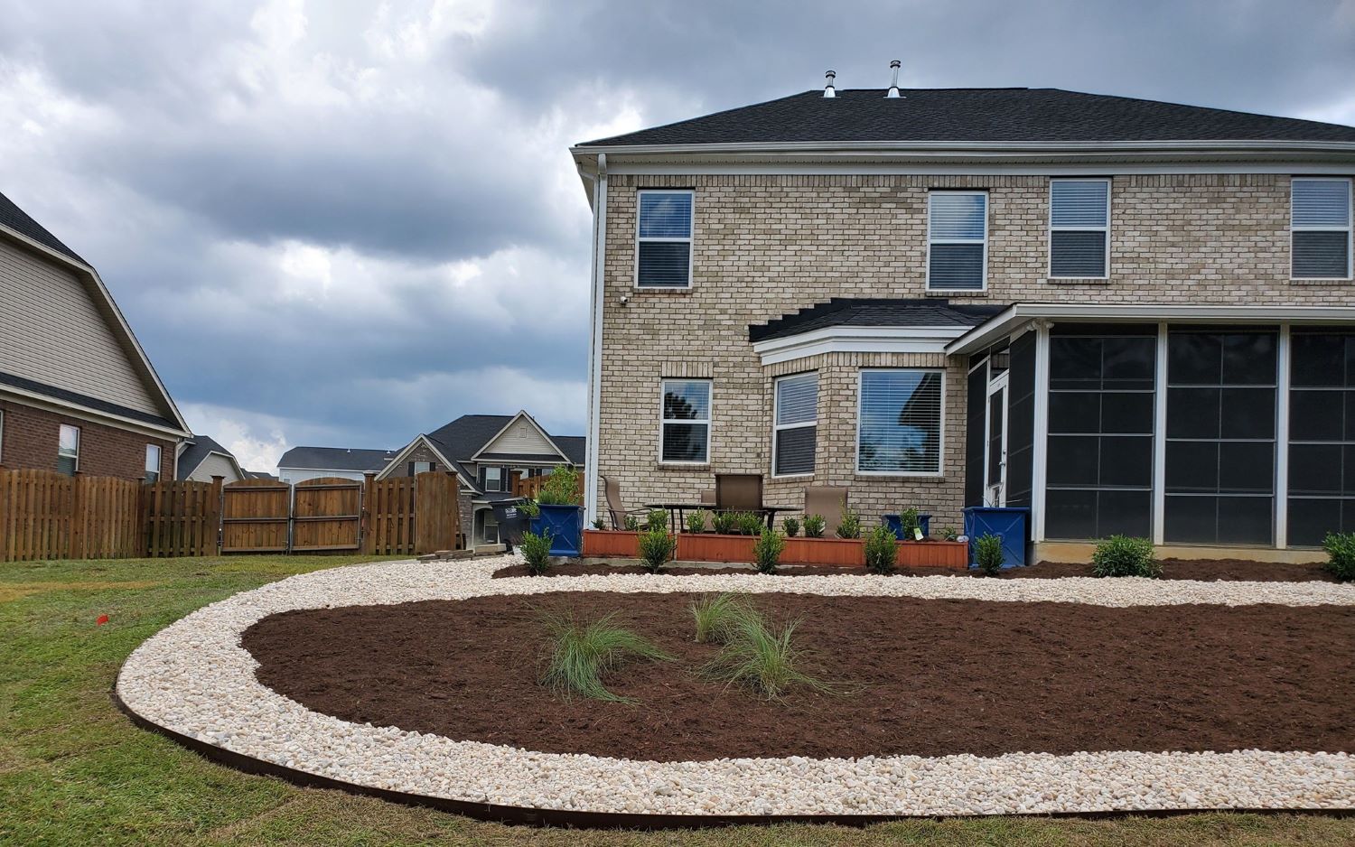 Backyard of a modern brick house featuring a fresh mulch bed bordered with white decorative stones and a few newly planted shrubs.