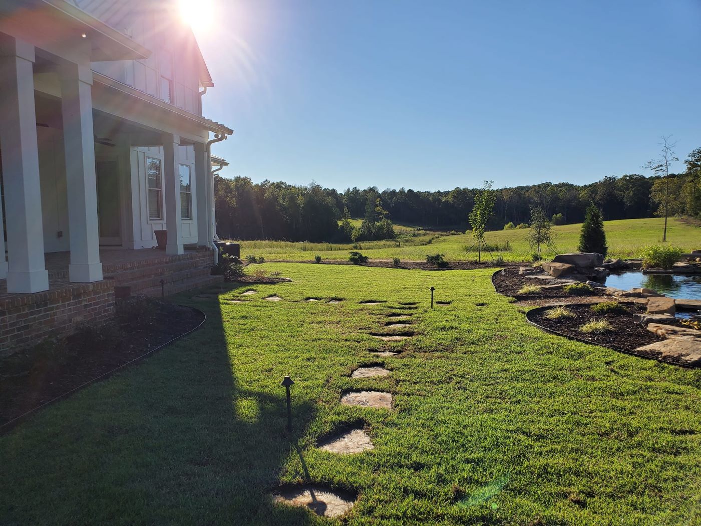 Stone pathway leading through a green lawn next to a house with scenic pond and trees in the background under a bright sun.
