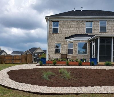 Backyard landscaping with fresh mulch bed bordered by white rocks behind a brick house.